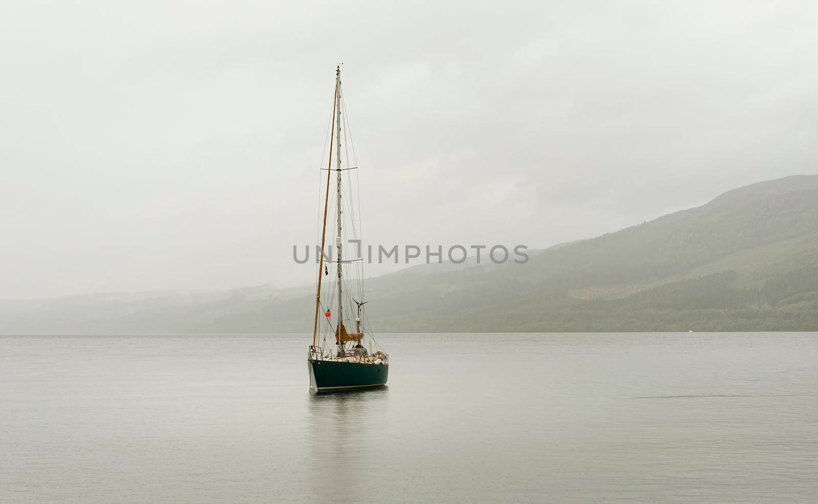 Landscape view of Loch Ness with calm water, mist and hills in the background and a sailboat with sails up floating peacefully on the water.