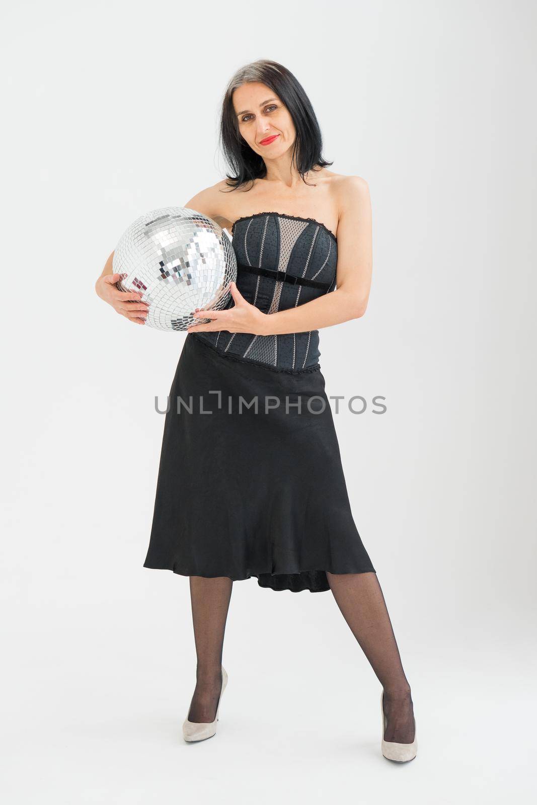 Studio photo of middle aged woman starting getting grey-haired wearing black clothes with silver disco ball in hands on white background, middle age sexy lady, happy life concept by balinska_lv