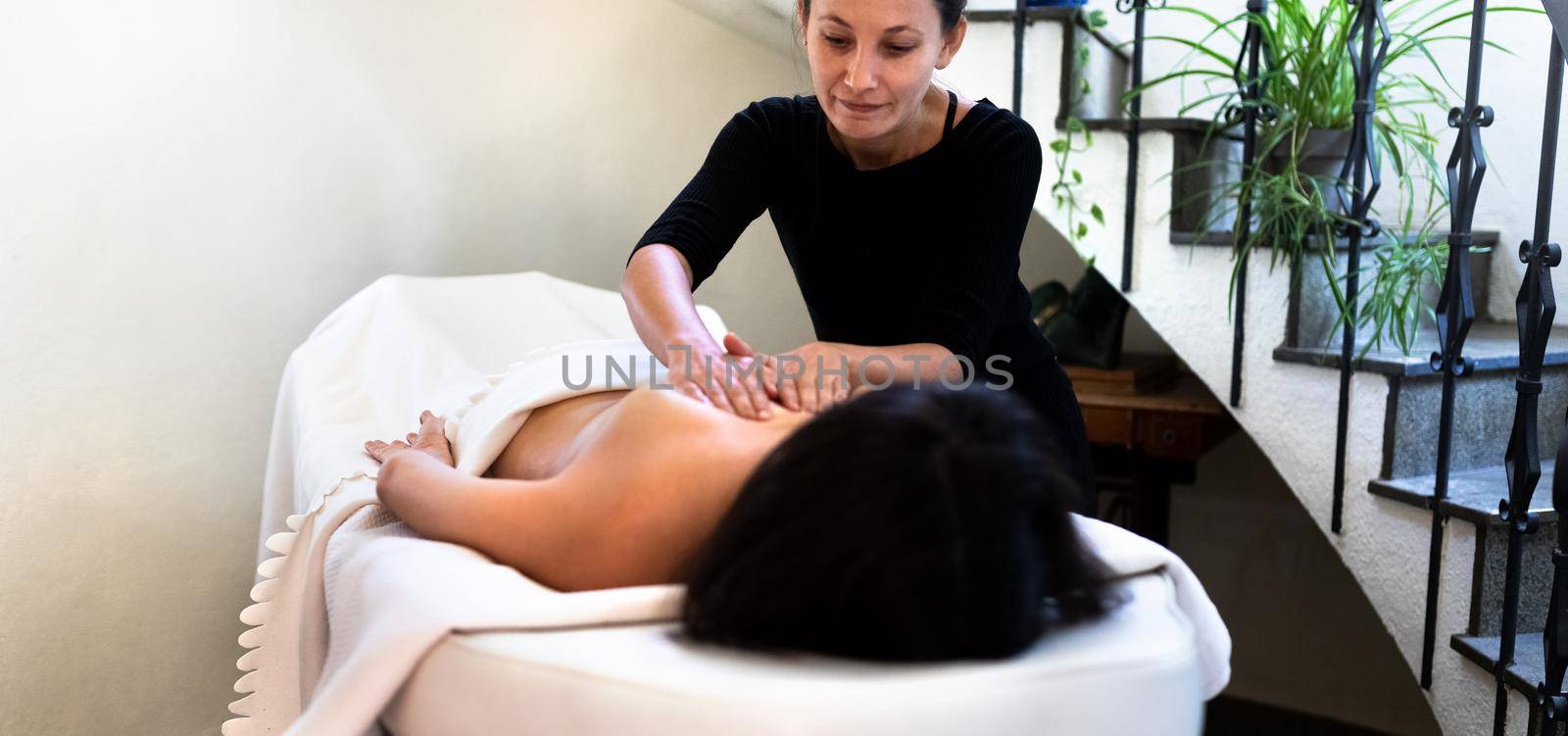 Panoramic image of latino female massage therapist giving back massage to caucasian woman. by Hoverstock