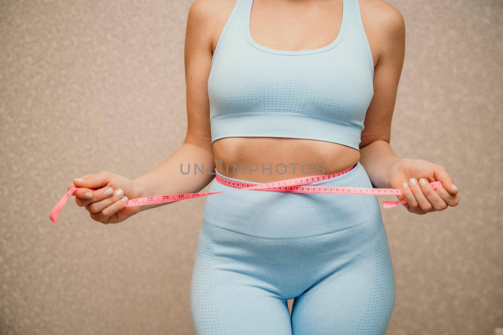 Cropped view of slim woman measuring waist with tape measure at home, close up. Unrecognizable european woman checking the result of diet for weight loss or liposuction indoors.