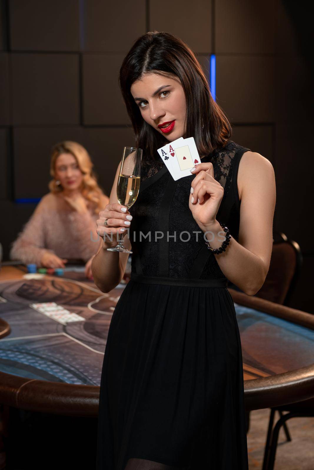 Confident gambling young brunette posing in casino on background of poker table, holding pair of aces and raising glass of champagne with winning look