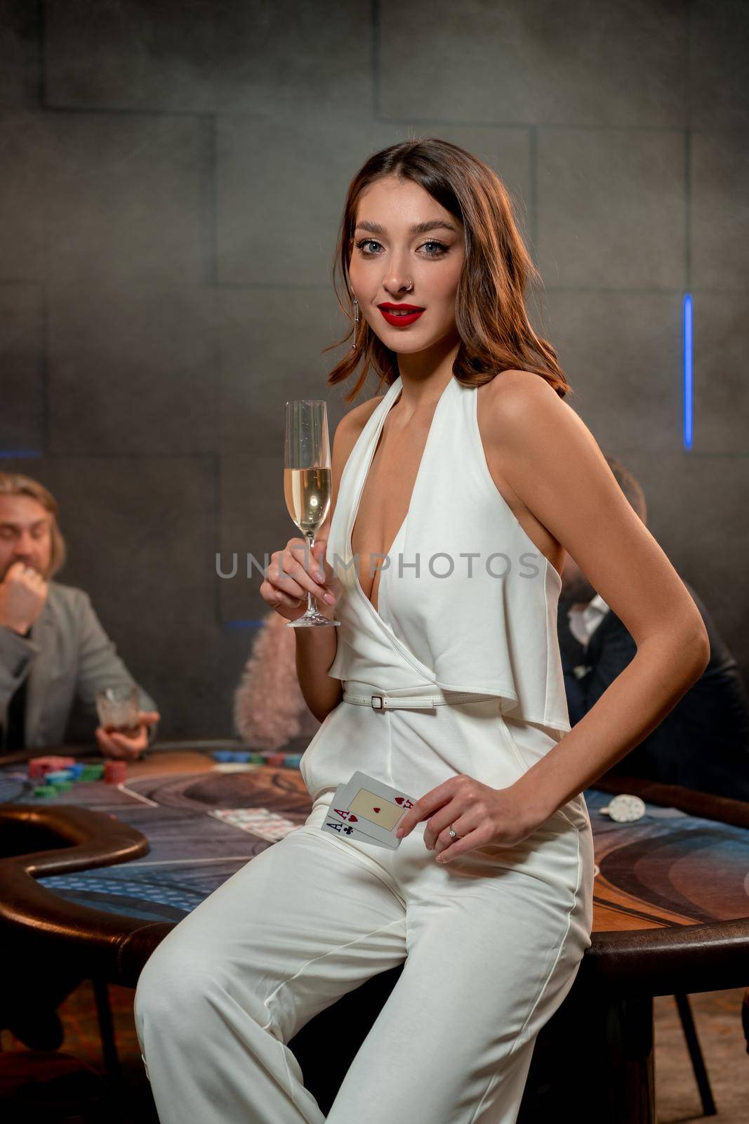Portrait of successful young female poker player sitting on edge of gaming table, holding winning pair of aces and raising glass of champagne