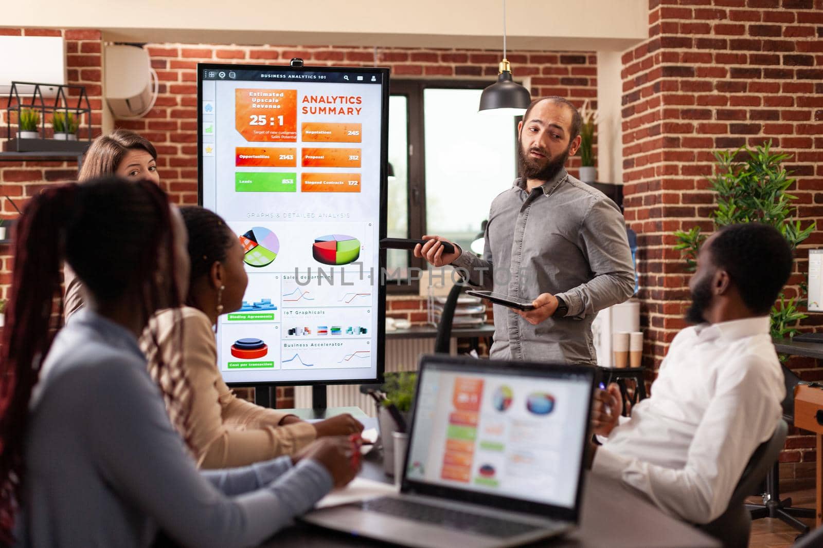 Businessman standing beside monitor showing management graphs explaining company strategy during business meeting in startup office. Diverse businesspeople working at financial presentation