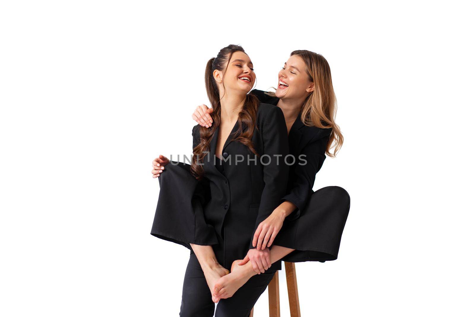 Two young business woman dressed black suit laugh and happy. One woman sitting chair and hugging other woman with barefoot legs. Positive caucasian female business people look at each other. Isolated