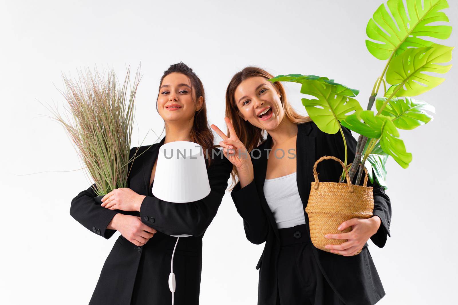 Two business woman dressed black suit smile holding houseplant and lamp standing isolated on white background. Moving concept. Relocation new office with positive. One woman shoving victory sign