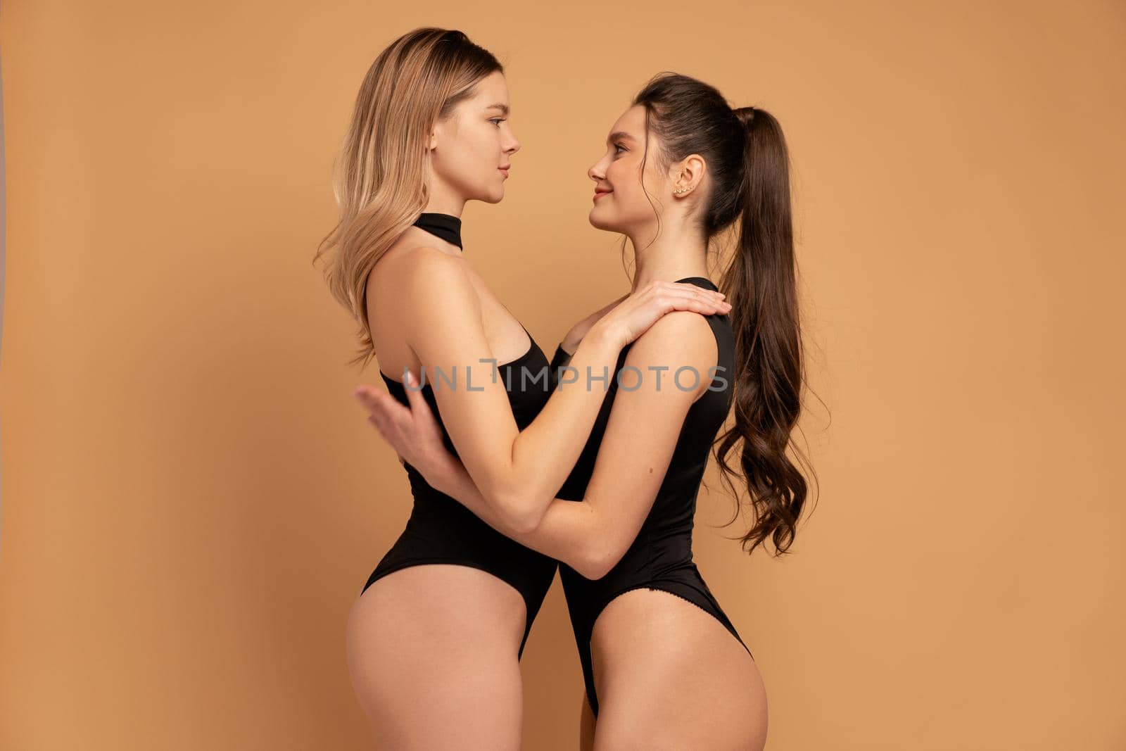 Two woman dressed black bodysuit embracing facing each other standing face to face isolated on color background. Beautiful female 20-25 years old studio shoot