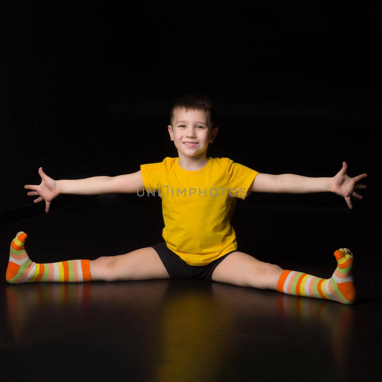 Boy sitting on floor with legs and hands wide apart. Happy smiling boy acrobat or gymnast in sportswear and striped socks performing stretching exercise against isolated black background in studio