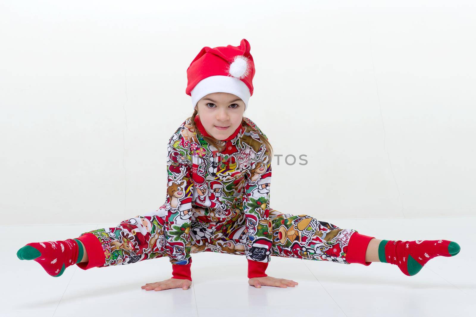 Happy little girl gymnast doing handstand and split. Cheerful flexible strong girl wearing colorful overalls and Santa Claus cap performing splits against isolated white background