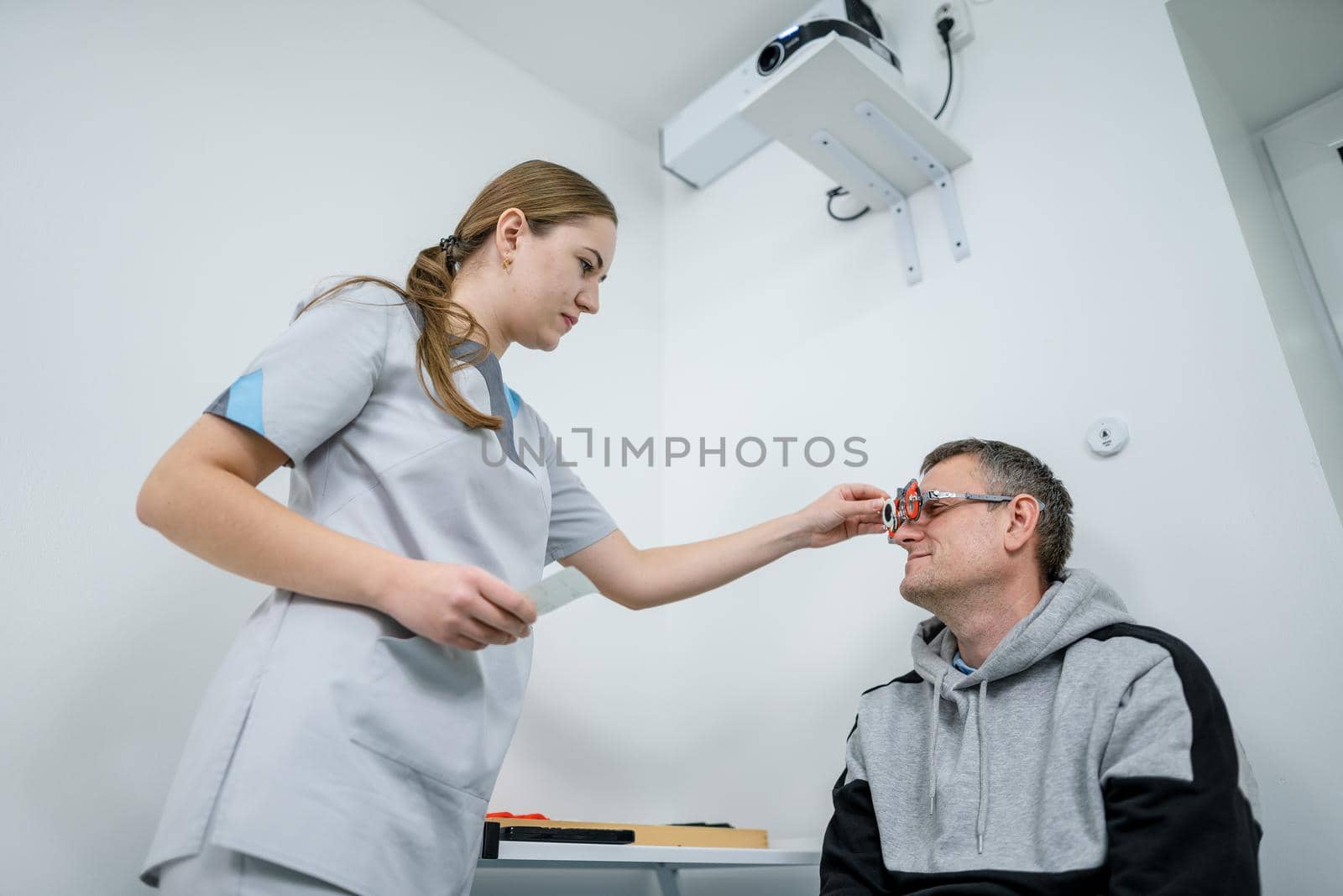 Vision correction. Selection of eyeglasses. Professional trial frames on male patient face while doctor checks eyesight in modern ophthalmologist office. Optometrist checking prescription for glasses by Tomashevska