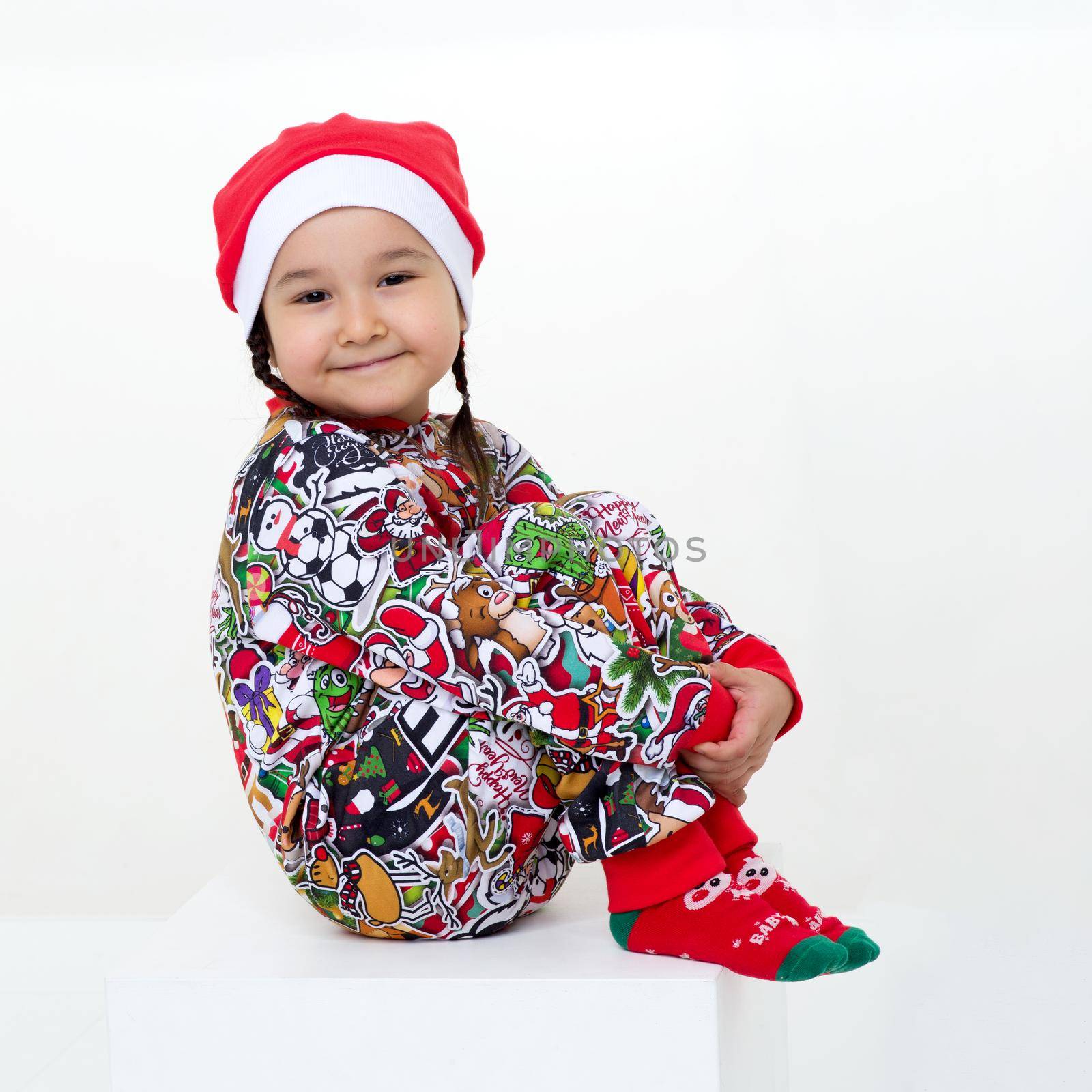 Happy girl in Christmas costume sitting on floor. Cute little girl wearing colorful overalls, red socks and Santa Claus cap sitting on floor hugging her knees against isolated white background