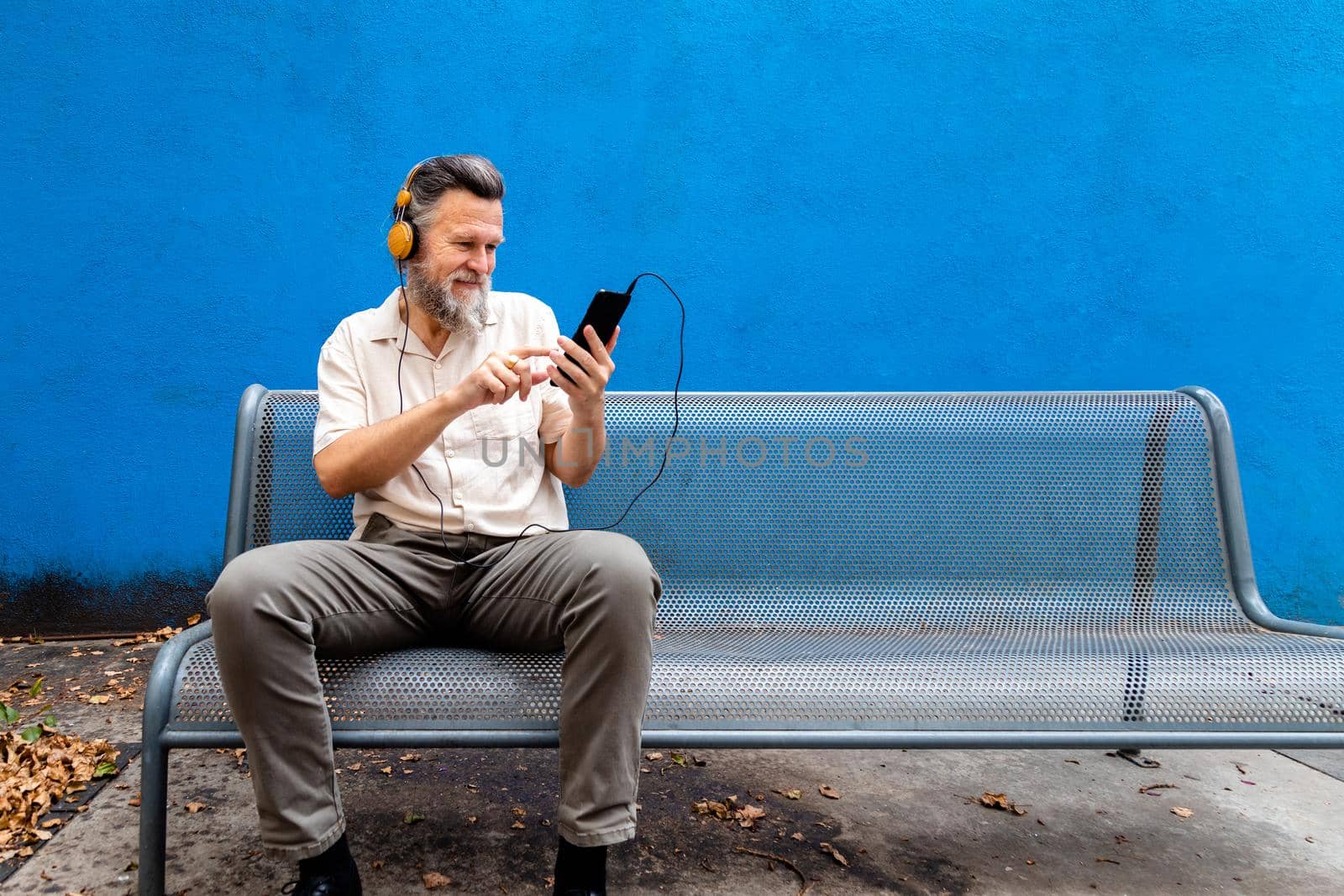 Mature caucasian man sit on a bench in public park using phone and listening to music with headphones. Copy space. Lifestyle concept.