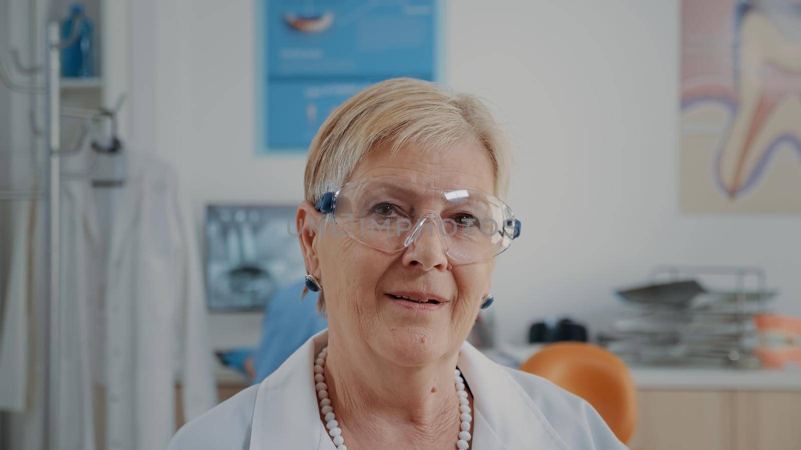 Portrait of senior dentist sitting in stomatology office, wearing protective glasses. Stomatologist preparing for dentistry appointment, working in oral care cabinet. Close up