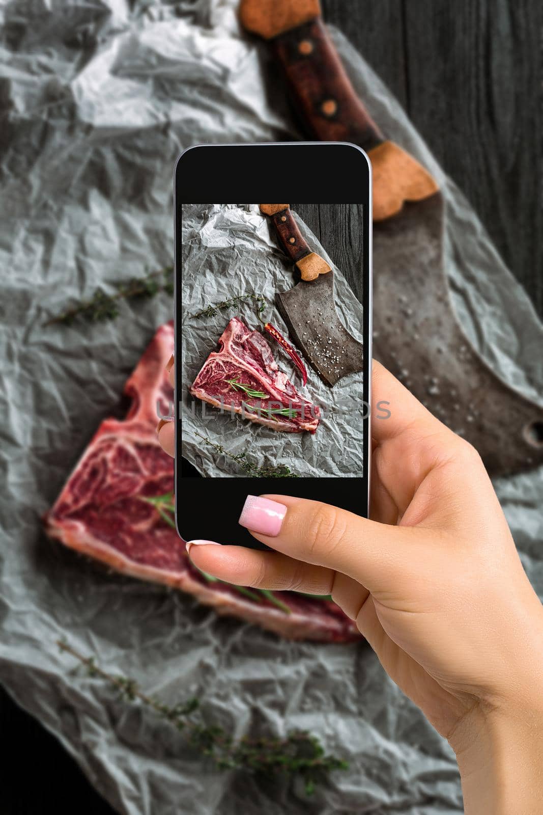 Photographing food concept - woman takes picture of raw dry aged t-bone steaks for grill with fresh herbs and cleaver by nazarovsergey