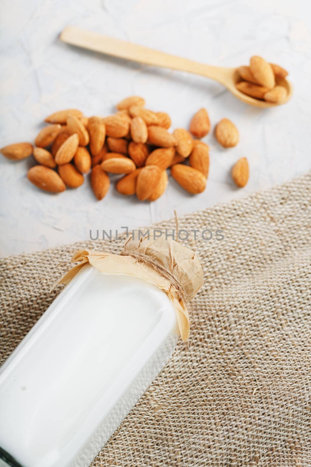 Milk from organic almonds in a transparent bottle with a scattering of seeds and a wooden spoon on a light background. Top view