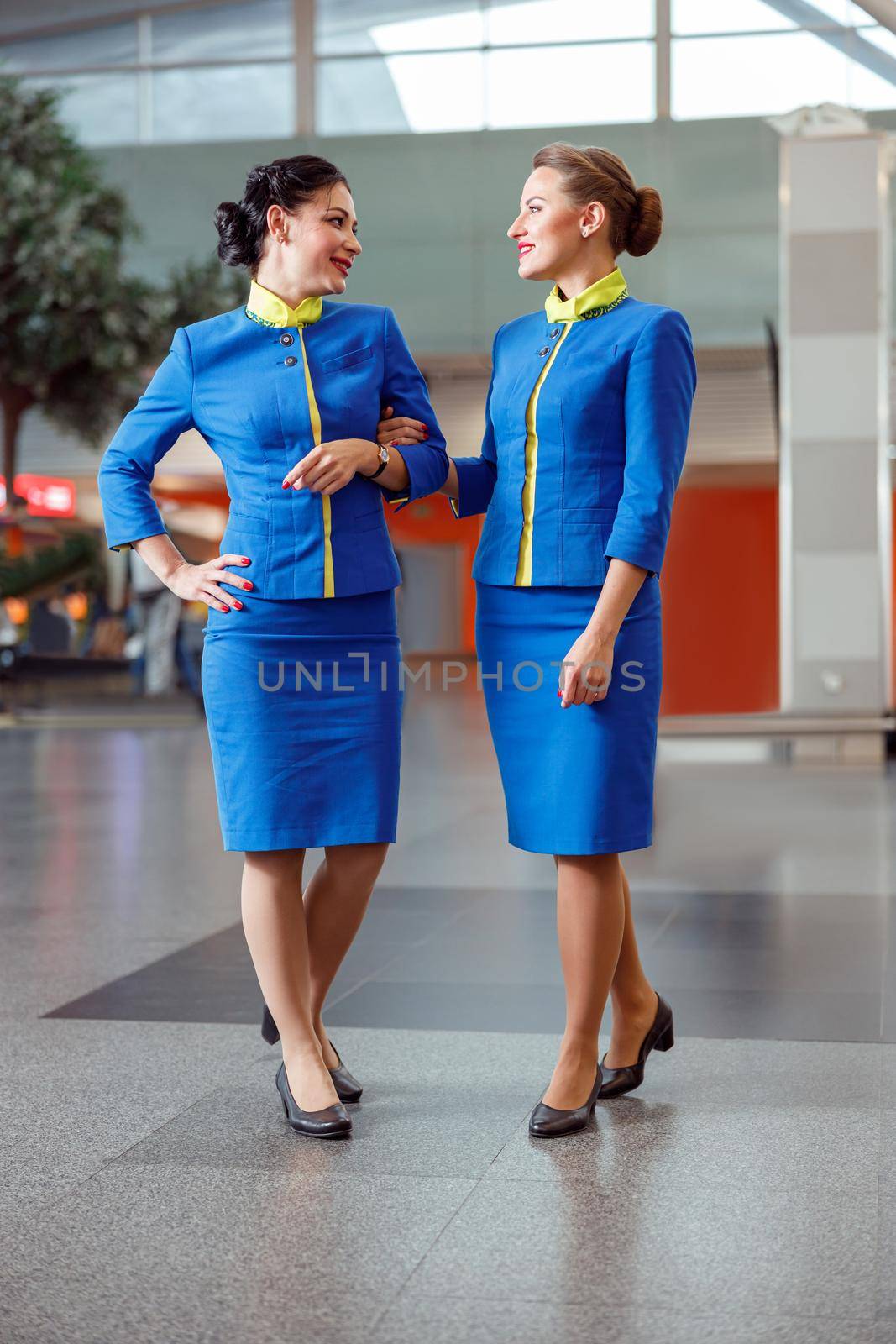 Full length of joyful woman flight attendant in aviation uniform looking at air hostess colleague and smiling while holding her arm