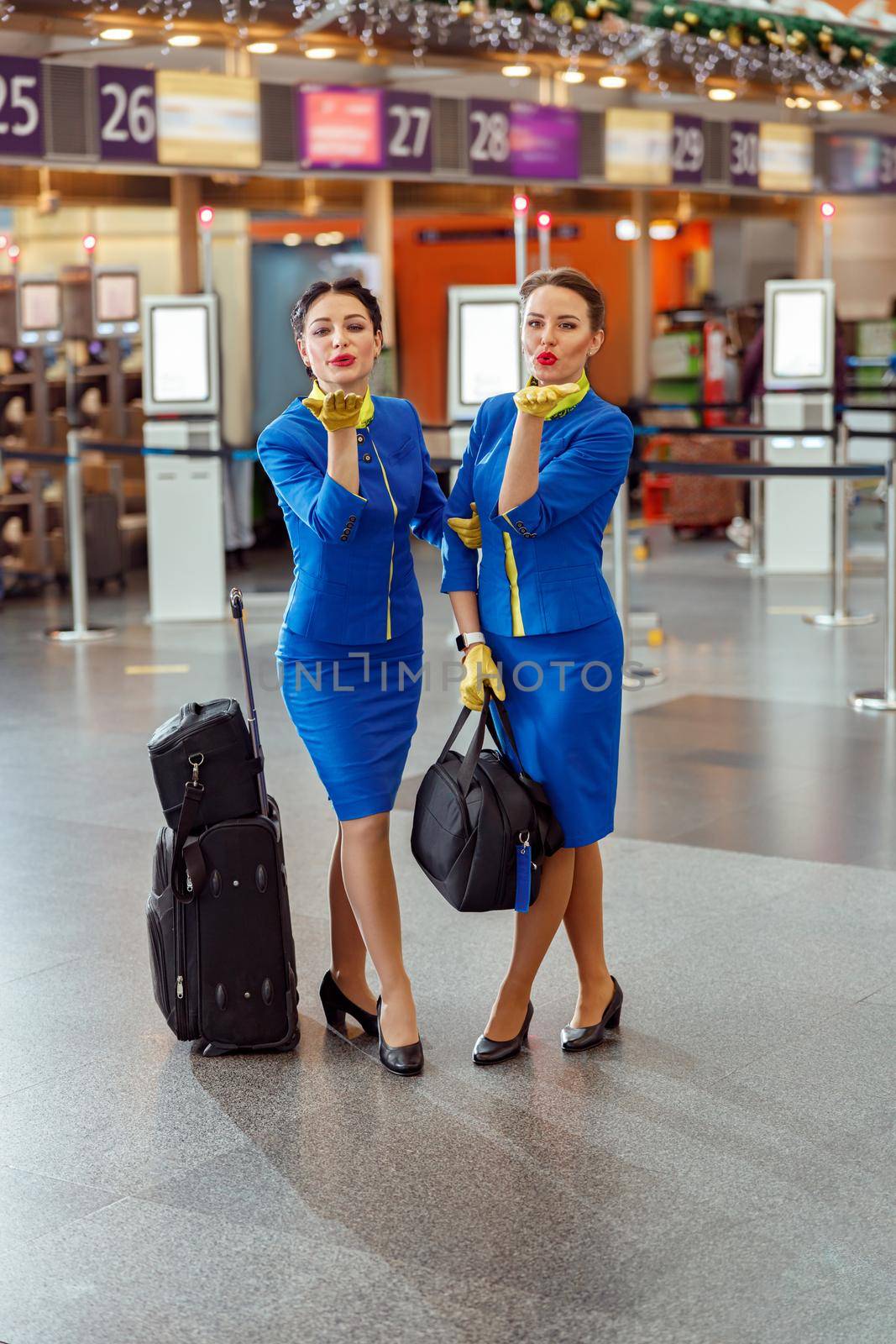 Full length of female flight attendants with travel bags looking at camera and sending air kiss while standing in passenger departure terminal