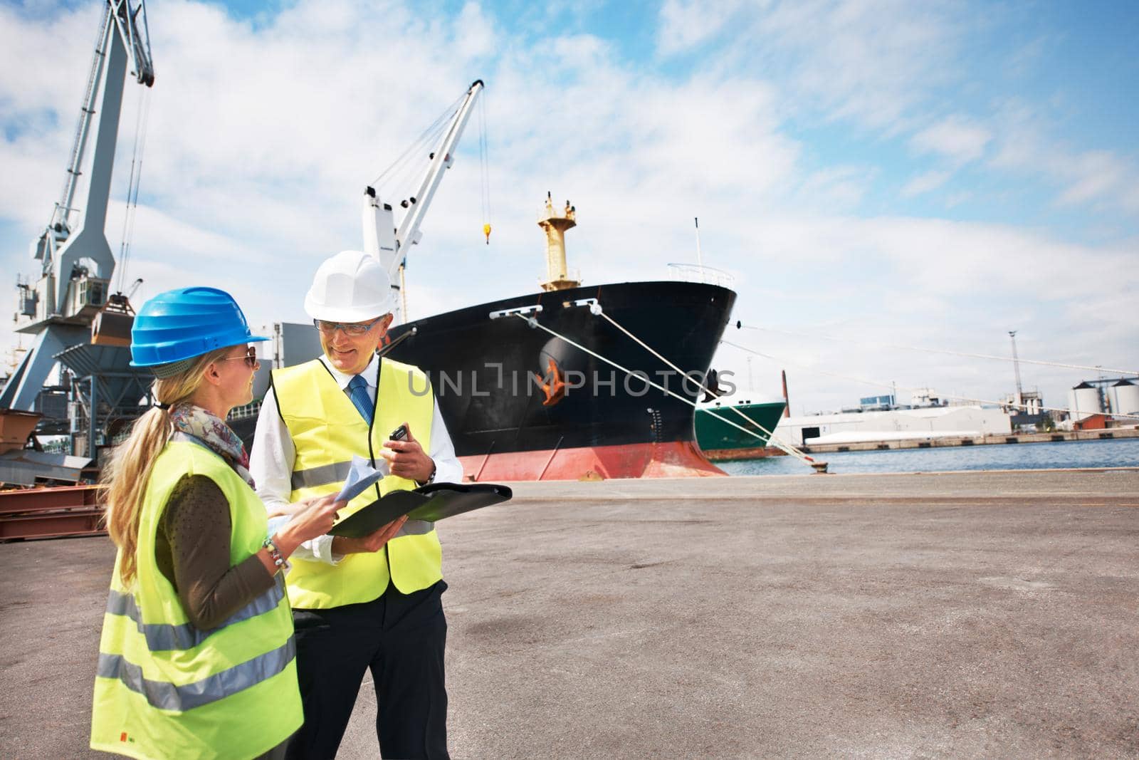 Open communication brings results. Two dock workers holding paperwork while standing in the shipyard. by YuriArcurs