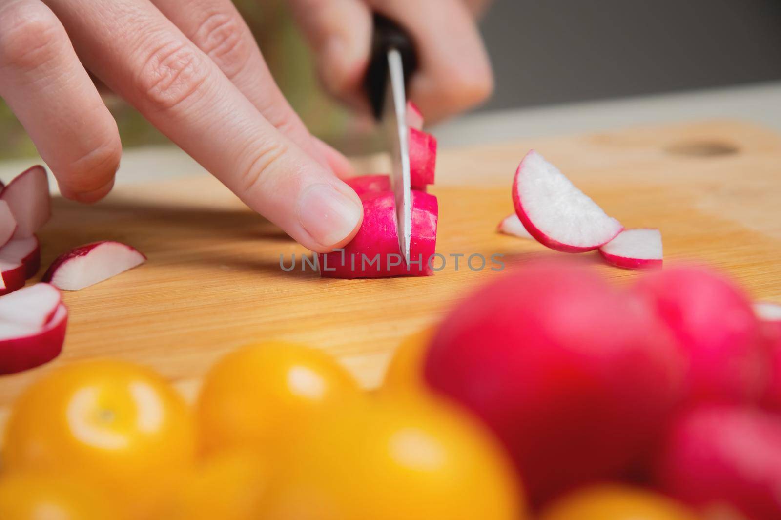 Close-up of female hands cut fresh raw radish on a wooden board against the background of yellow cherry tomatoes. Vegetarian tasty and healthy food by yanik88