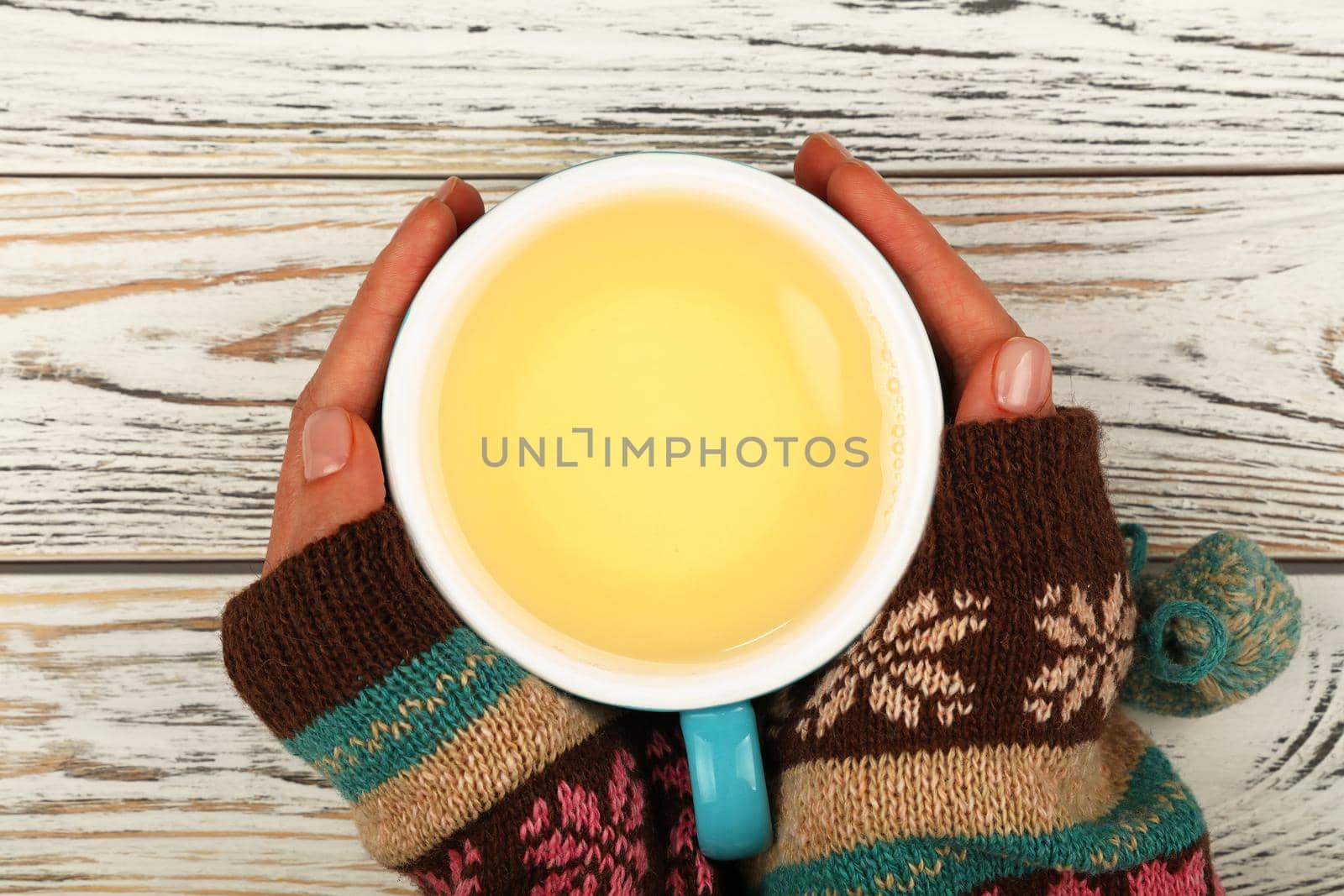 Close up two woman hands hold and hug big full cup of green oolong tea over white wooden table, elevated top view, directly above