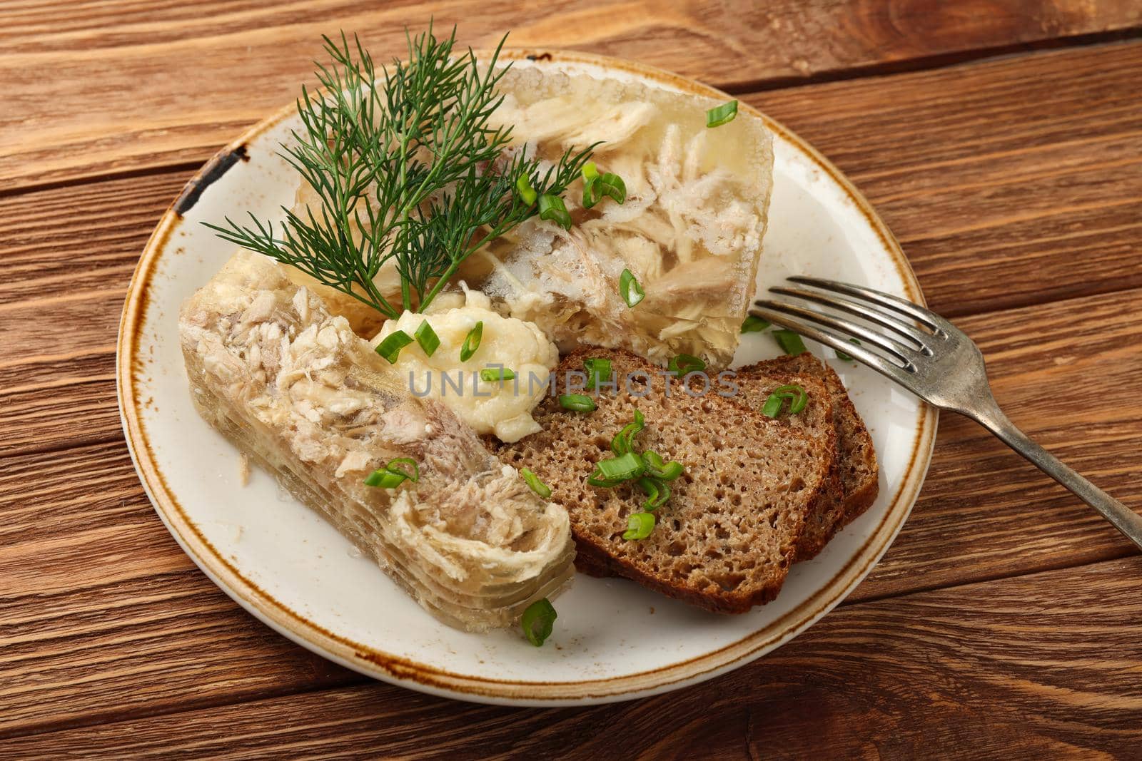 Close up portion of aspic or traditional Eastern European chicken meat jelly with slices of rye bread and horseradish dressing sauce, on brown wooden table, high angle view