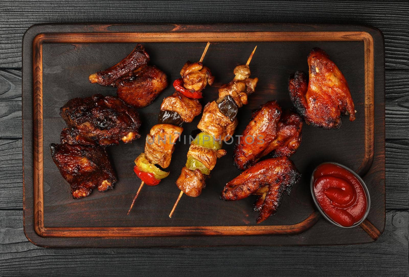 Close up portion of mixed grilled barbecue combo plate with chicken buffalo wings, beef spare ribs and skewer meat on wooden board over black table planks, elevated top view, directly above