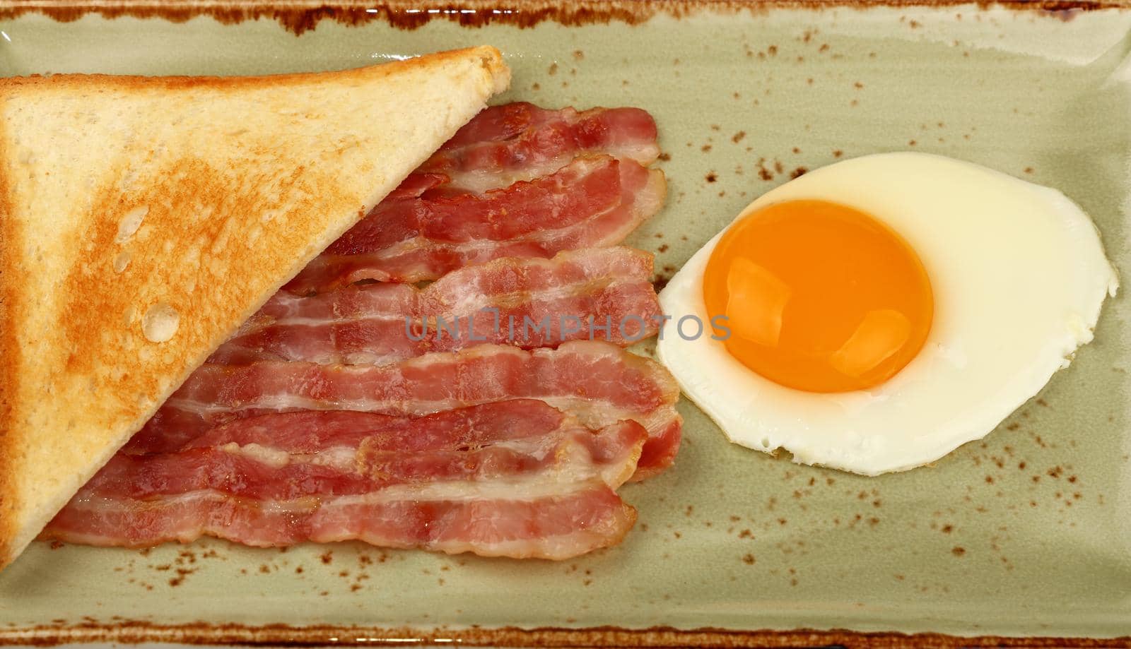 Close up English breakfast, sunny side egg, half of toasted bread and roasted bacon slices on blue plate, high angle view