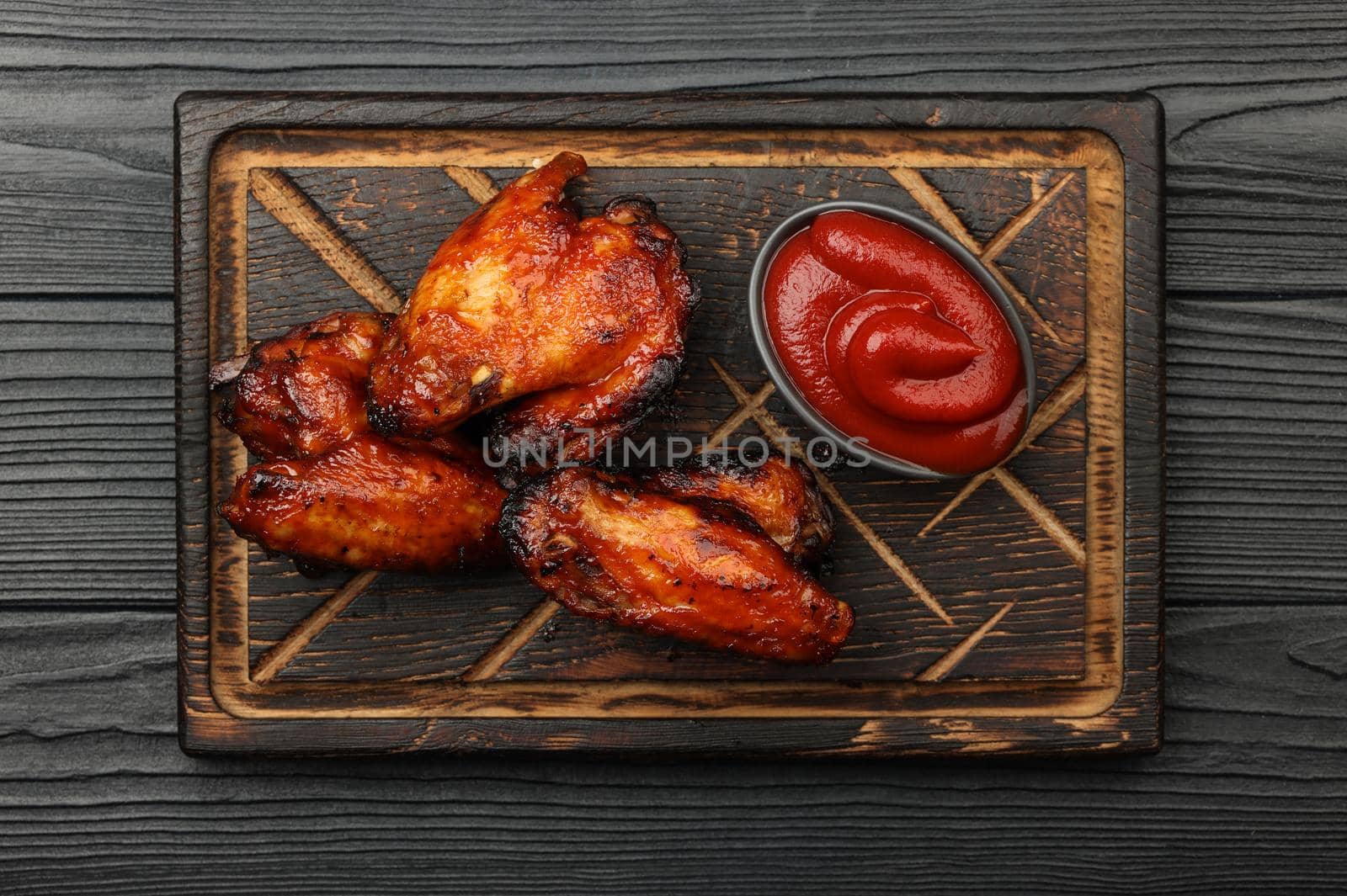 Close up portion of grilled barbecue chicken buffalo wings and BBQ tomato sauce served on wooden board over black table planks, elevated top view, directly above