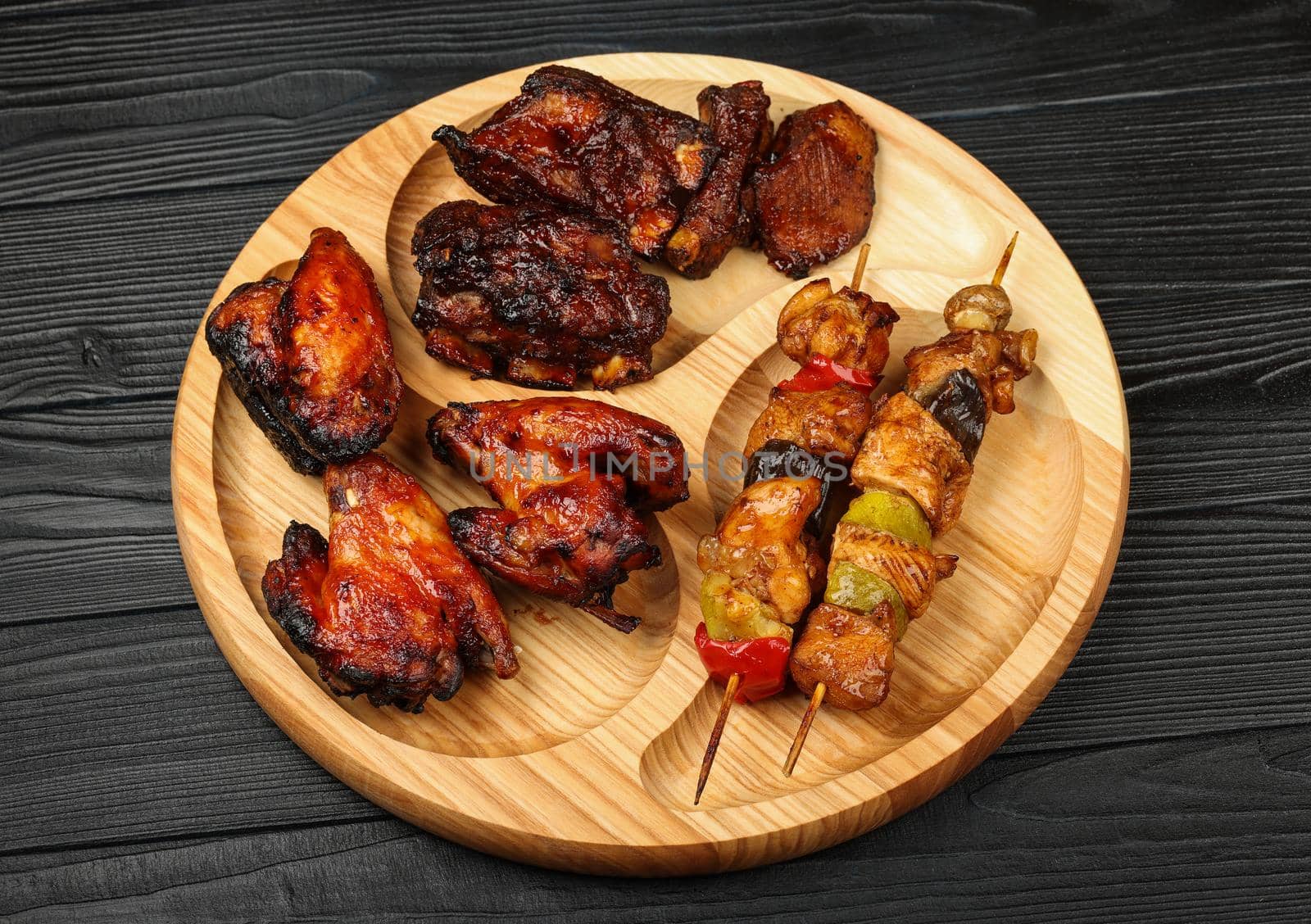 Close up portion of mixed grilled barbecue combo plate with chicken buffalo wings, beef spare ribs and skewer meat on wooden board over black table planks, high angle view