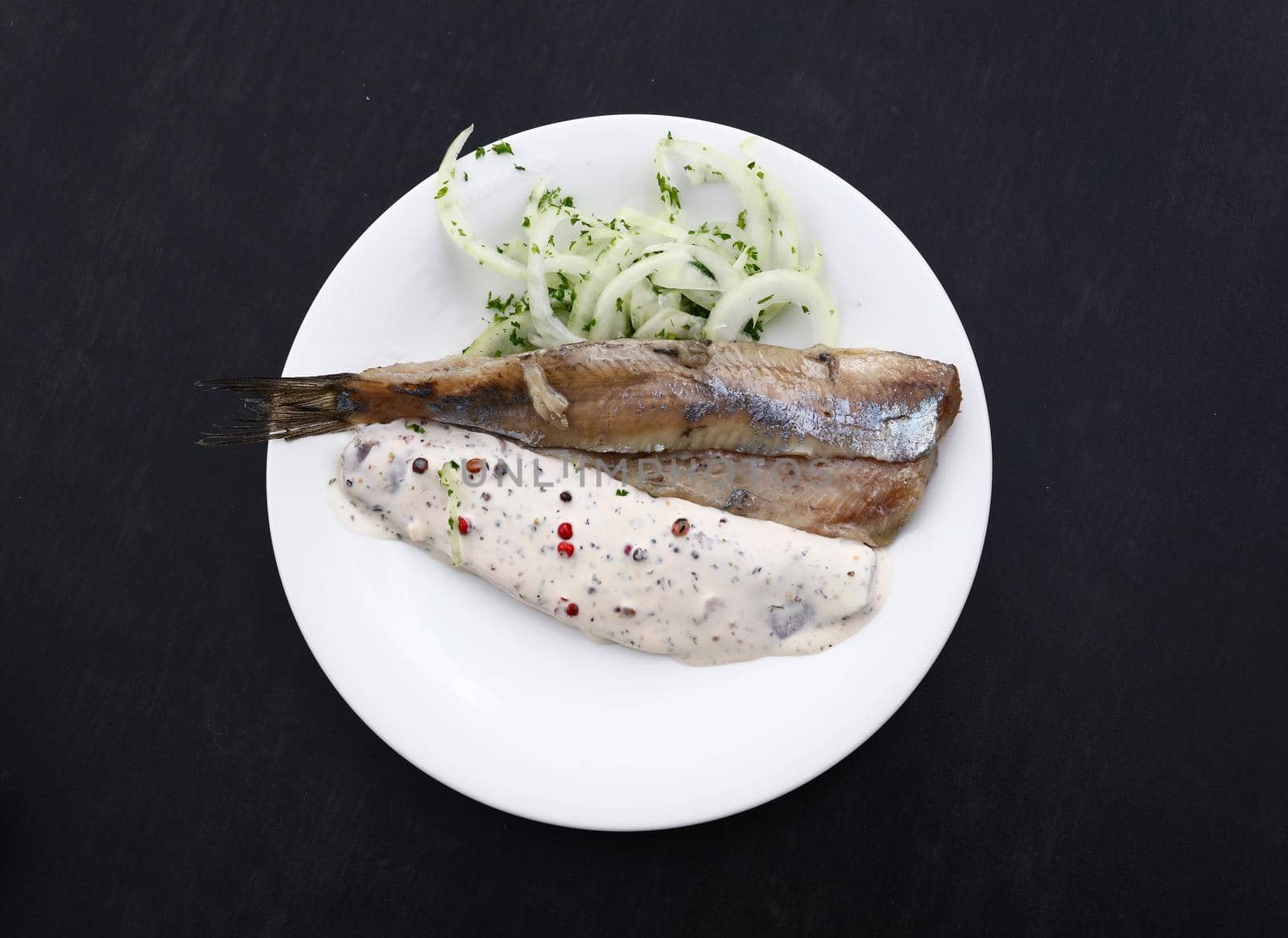 Close up portion of salted pickled herring fish filets with onions and sauce on white plate over black table, elevated top view, directly above