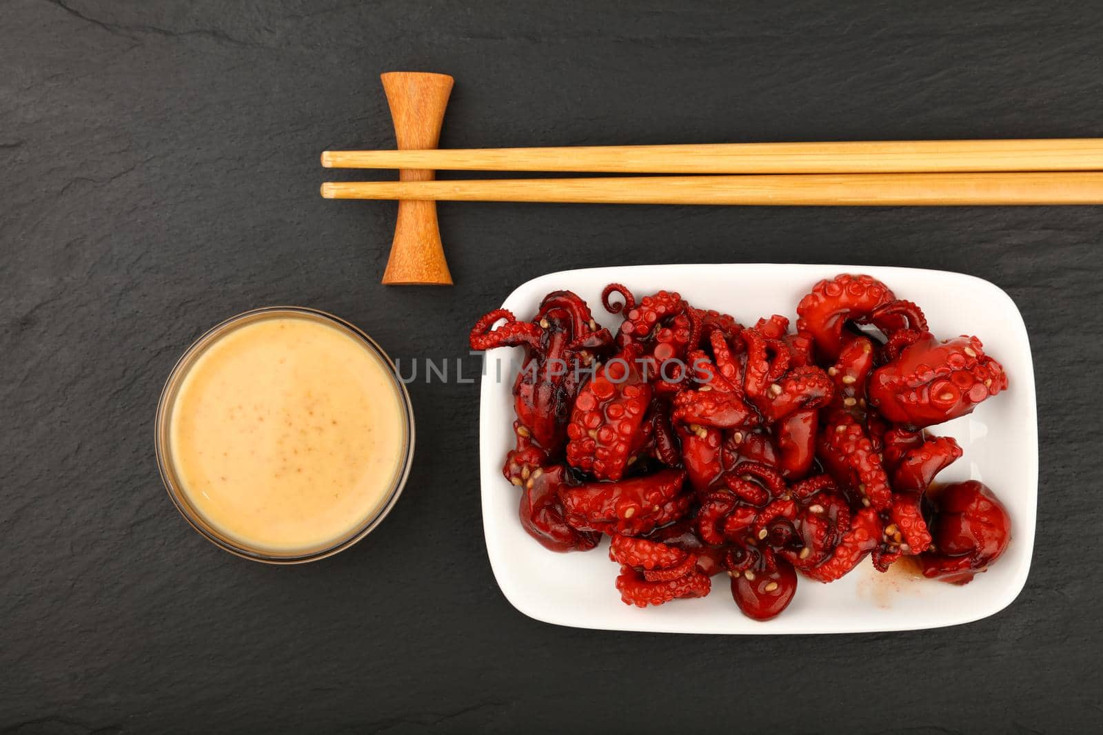 Close up portion of seafood small whole red baby octopus salad with satay sauce and chopsticks on black slate board background, elevated top view, directly above