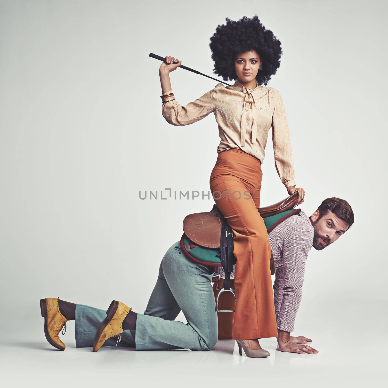 A studio shot of an attractive woman in 70s wear riding a handsome man wearing a saddle while using a riding crop