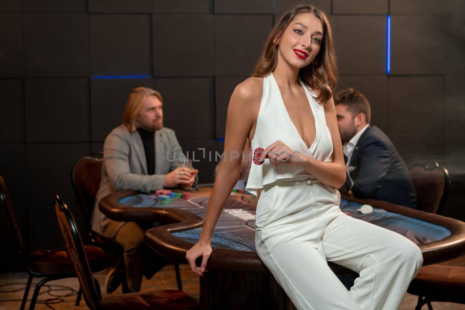 Portrait of smiling attractive young woman in stylish white jumpsuit sitting on edge of poker table in gambling establishment room with other players in background, holding betting chips