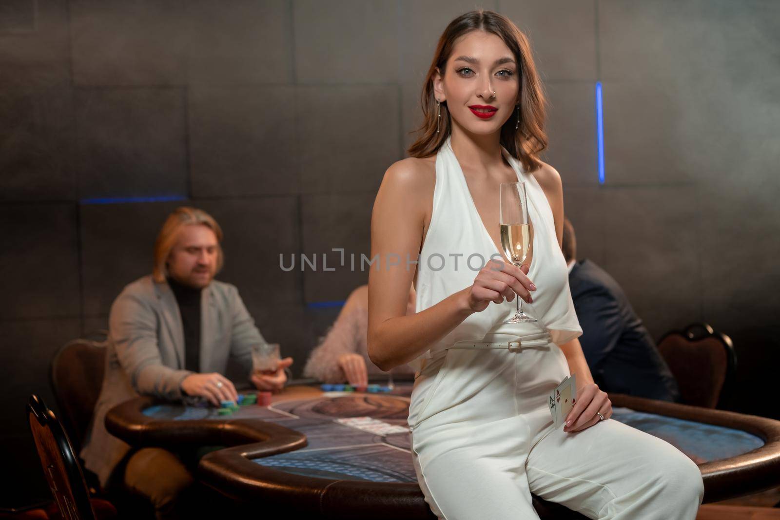 Portrait of young brown-haired woman wearing elegant white jumpsuit, holding glass of champagne and cards in hands, posing in casino near poker table against background of players making bets