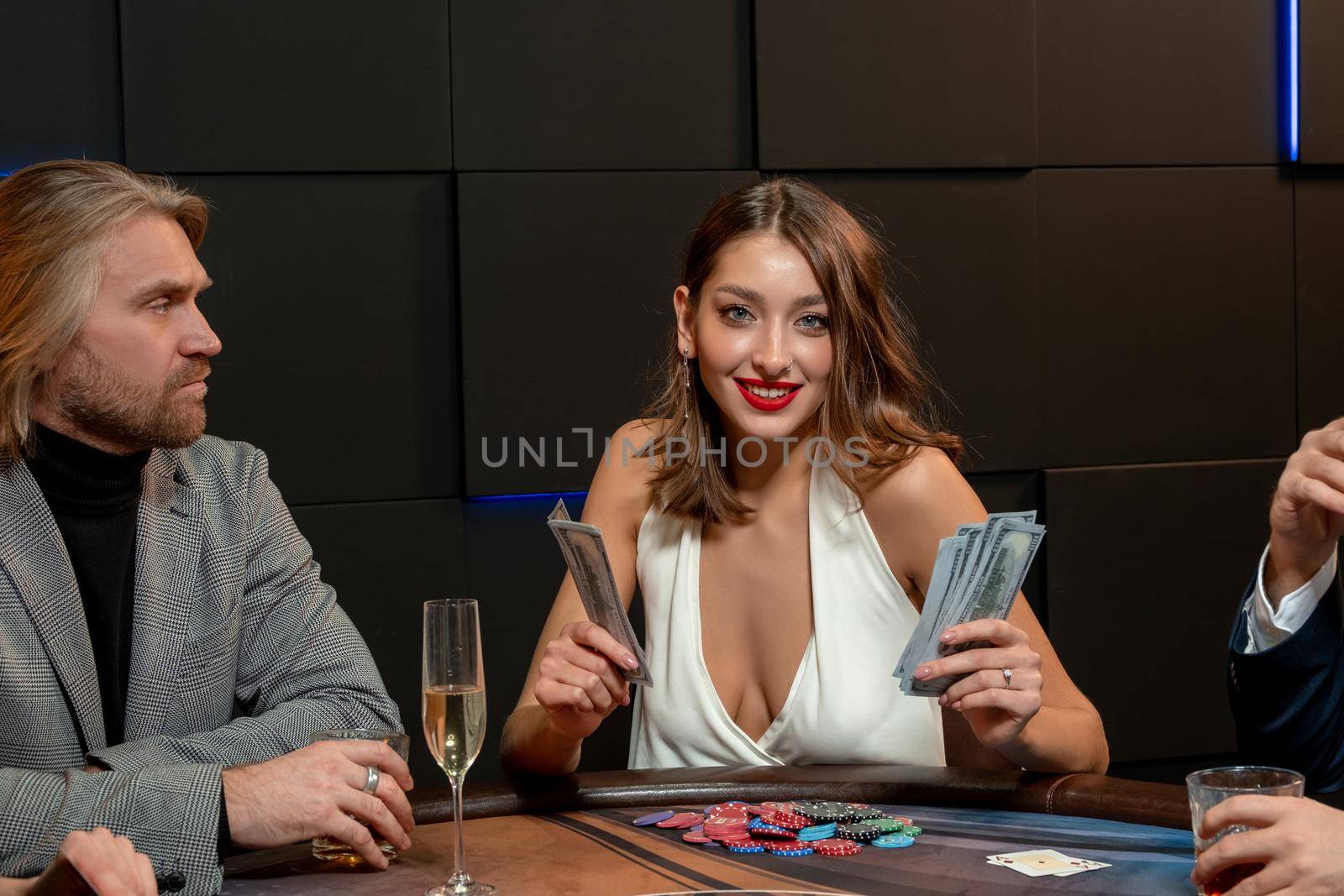 Cheerful stylish brown-haired girl breaking bank in poker game, counting banknotes while sitting at gaming table with opponents. Good luck gambling concept