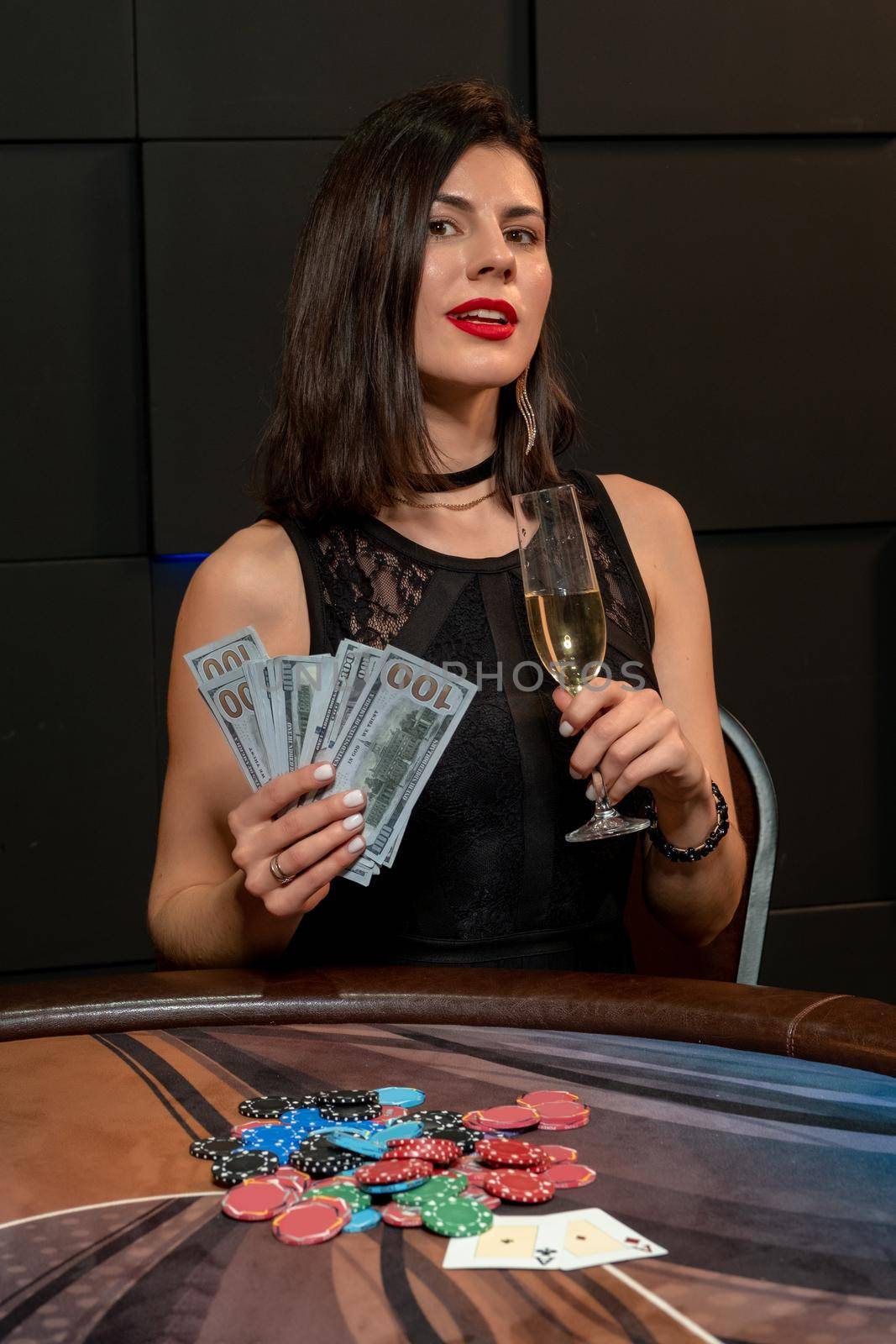 Lucky young brunette woman winning in poker, sitting at gaming table in casino with pile of chips lying in front of her, holding glass of champagne and fan of banknotes. Successful gambling concept