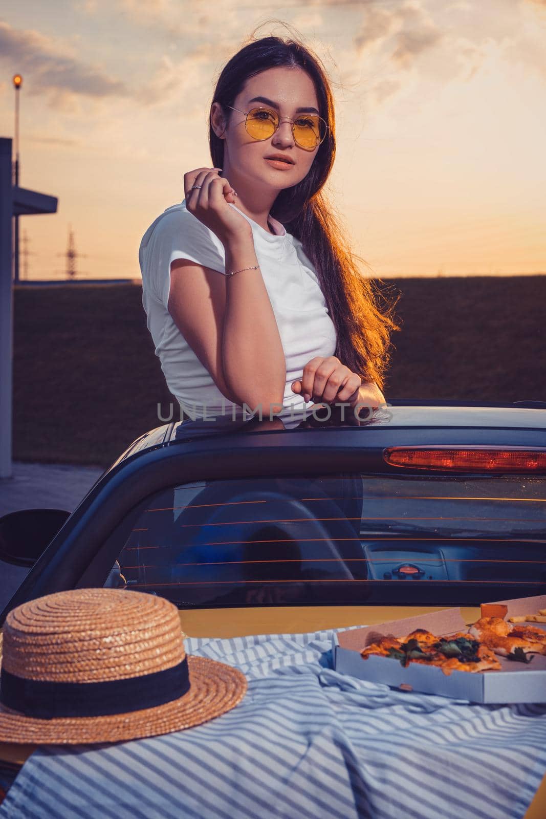 Young gorgeous woman in sunglasses, white t-shirt is posing in yellow car roadster with pizza, striped cloth and hat on its trunk. Fast food, unhealthy nutrition. Summer evening. Close up