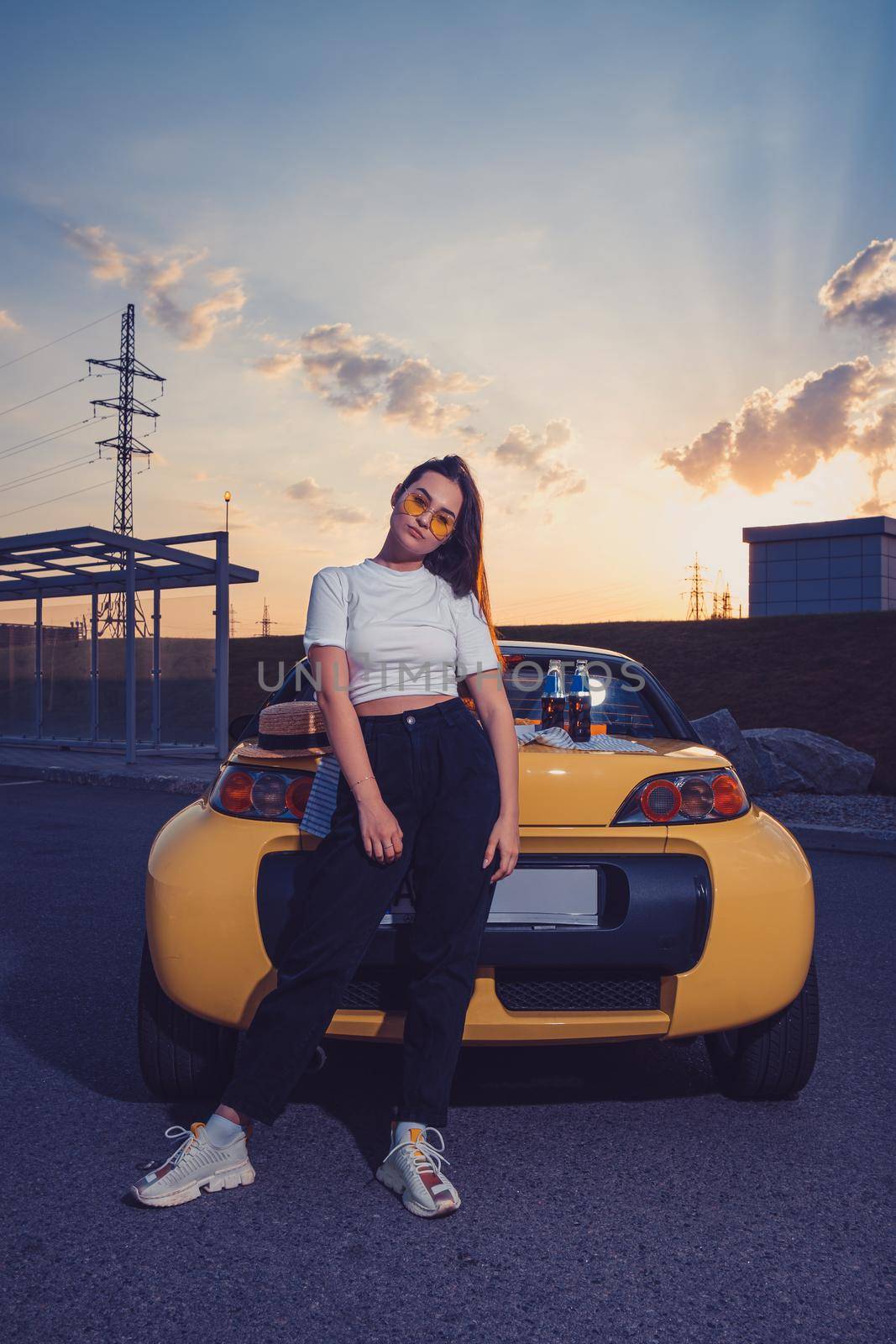Gorgeous young woman in sunglasses, white top, black pants and sneakers is posing outdoors near yellow car cabriolet with two glass bottles of soda on its trunk. Summer sunset. Full length, mock up