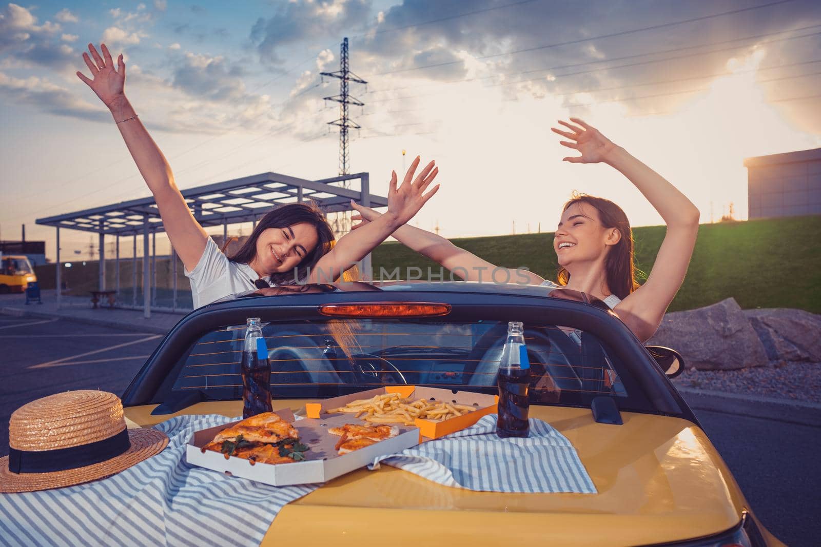 Young girls smiling, raised up hands, posing in yellow car with french fries, pizza and soda in glass bottles on its trunk. Fast food. Copy space by nazarovsergey