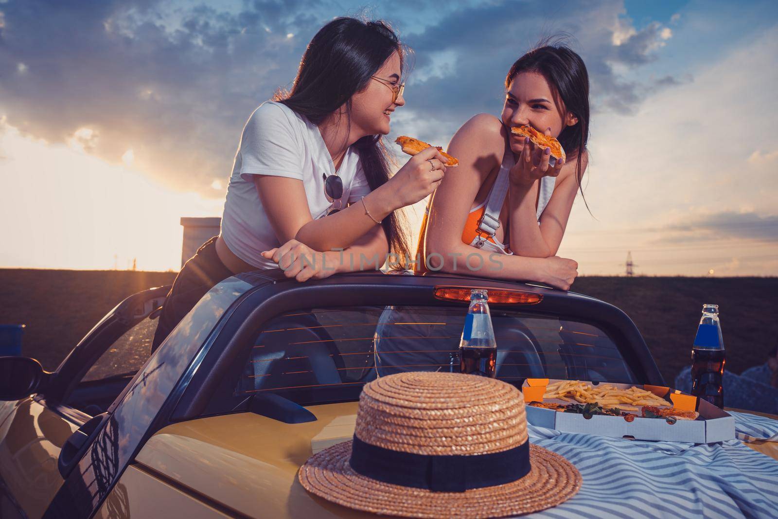 Two young girls are eating pizza, laughing, posing in yellow car with french fries, hat and soda in glass bottles on trunk. Fast food. Mock up by nazarovsergey