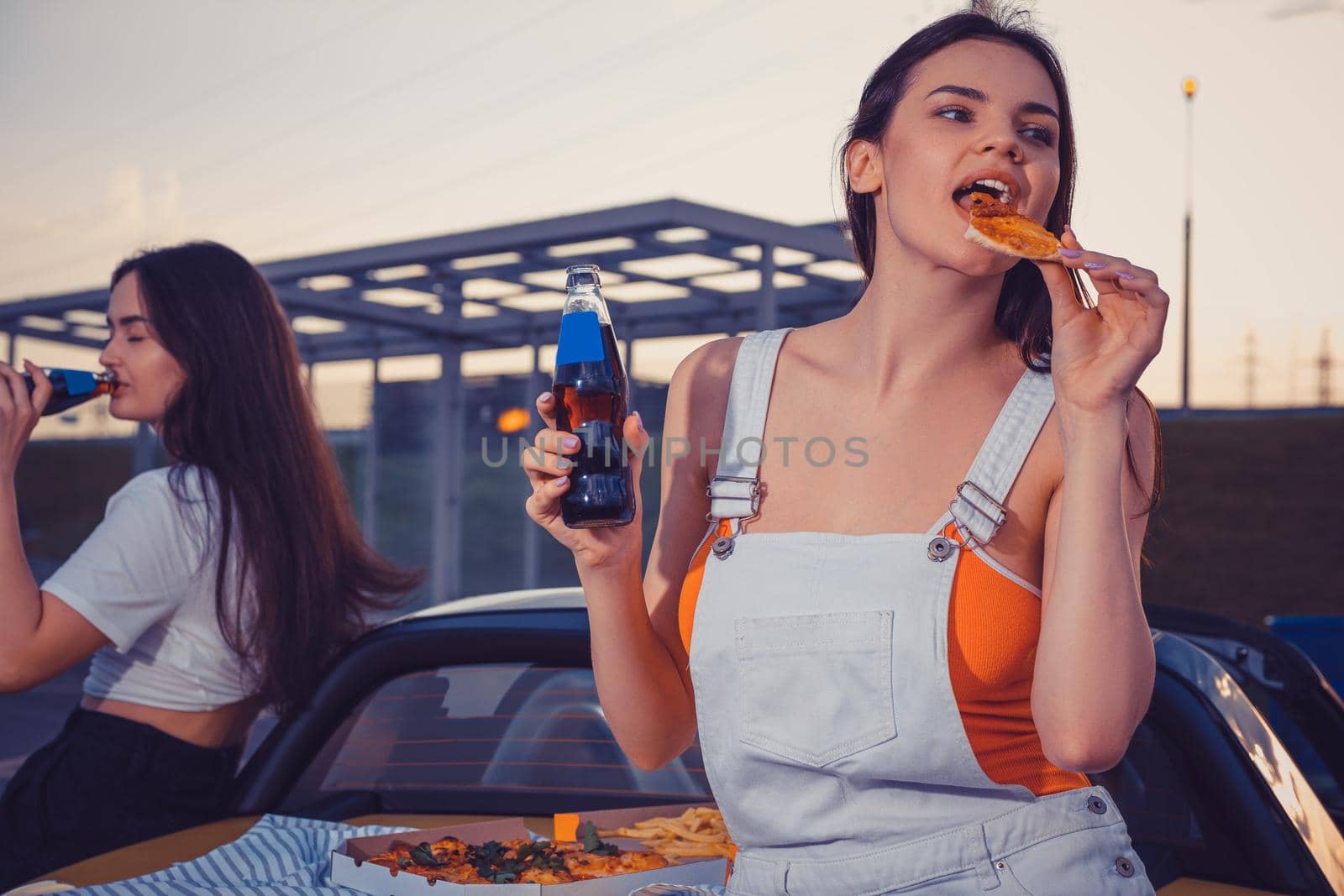 Attractive girls in casual outfit eating pizza, enjoying carbonated drink in glass bottles while posing near yellow car on parking lot. Copy space by nazarovsergey