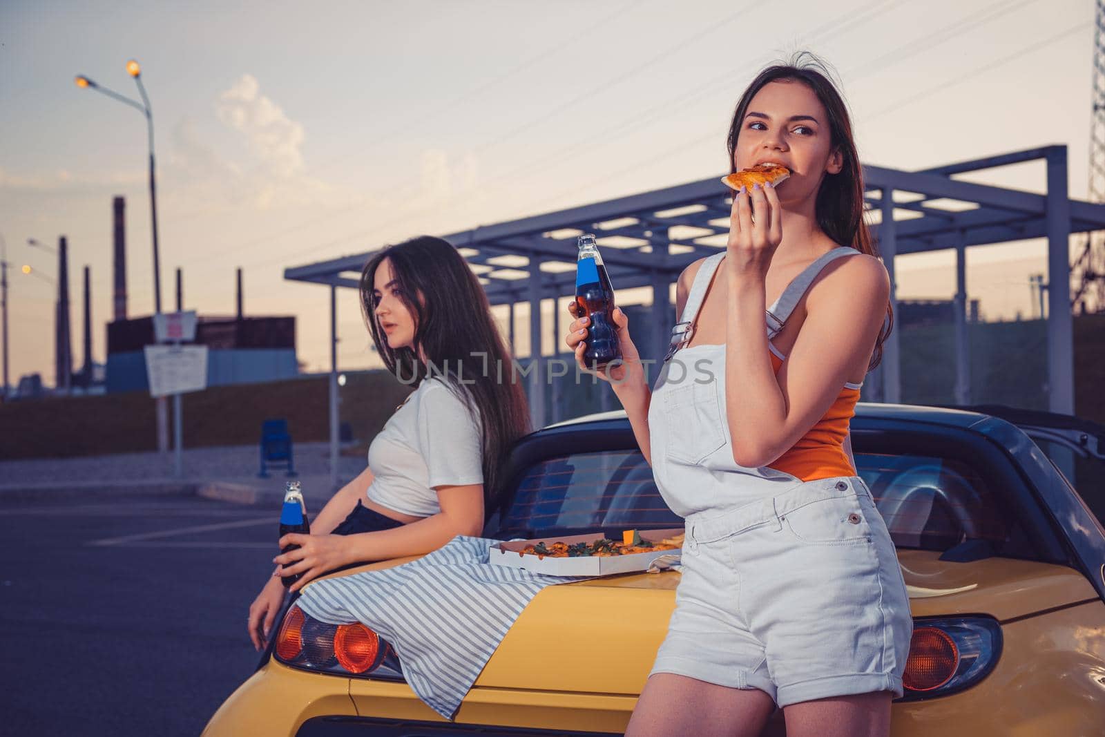 Attractive ladies in casual clothes eating pizza, enjoying soda water in glass bottles, posing near yellow car on parking lot. Close up, copy space by nazarovsergey