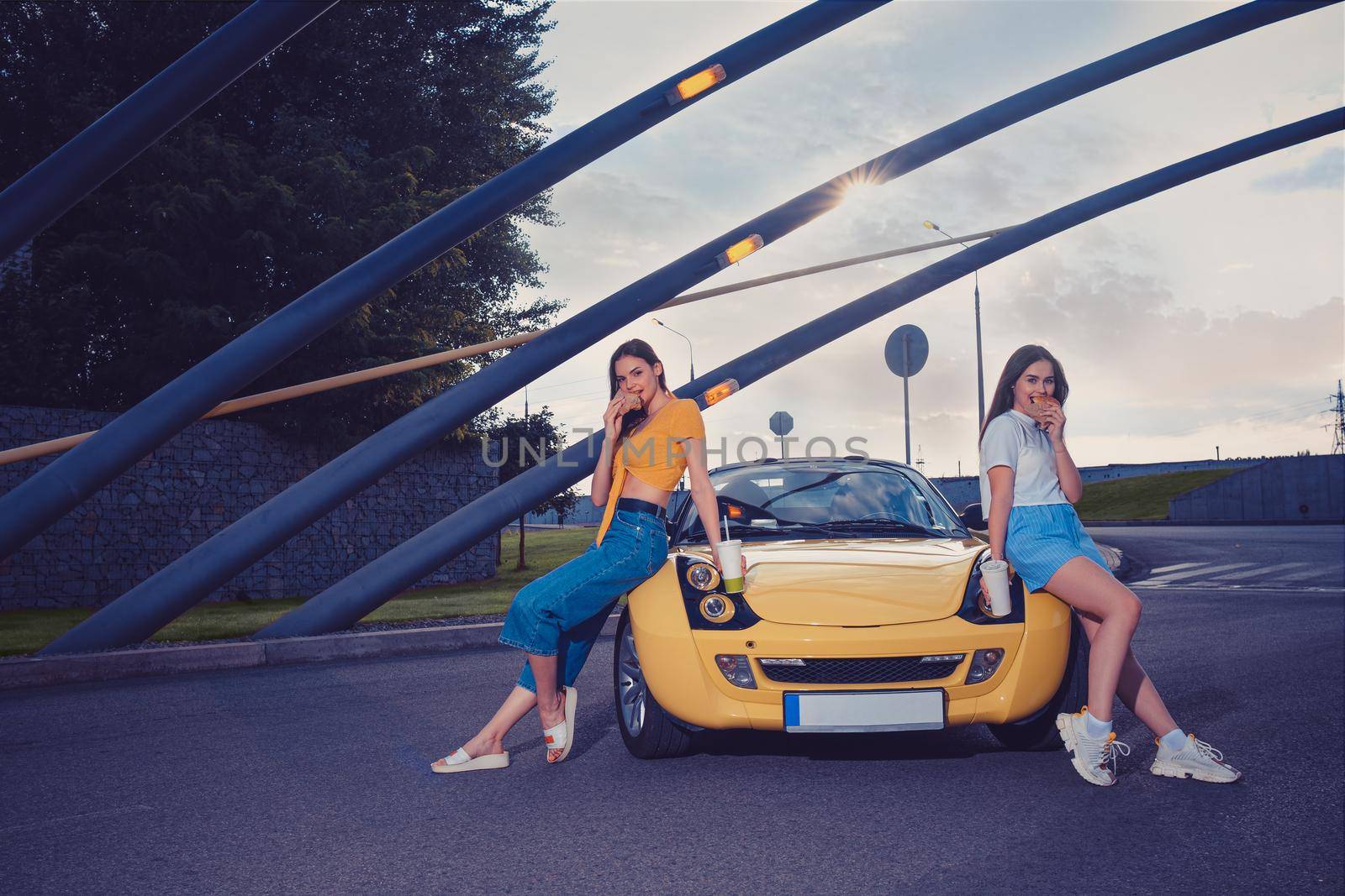 Girls in casual outfit eating hamburgers and holding beverages in paper cups while leaning on the hood of yellow car roadster. Fast food. Copy space by nazarovsergey