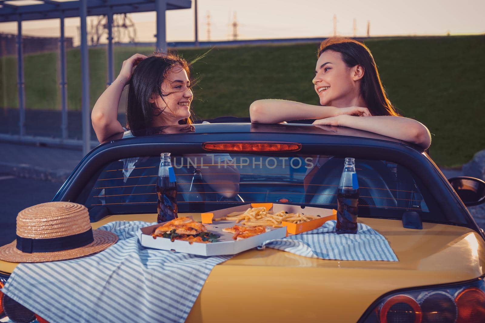 Pretty models in casual outfit are smiling, posing in yellow car cabrio with french fries, pizza, hat and soda in glass bottles on its trunk. Fast food. Summer evening. Close up, copy space, mock up