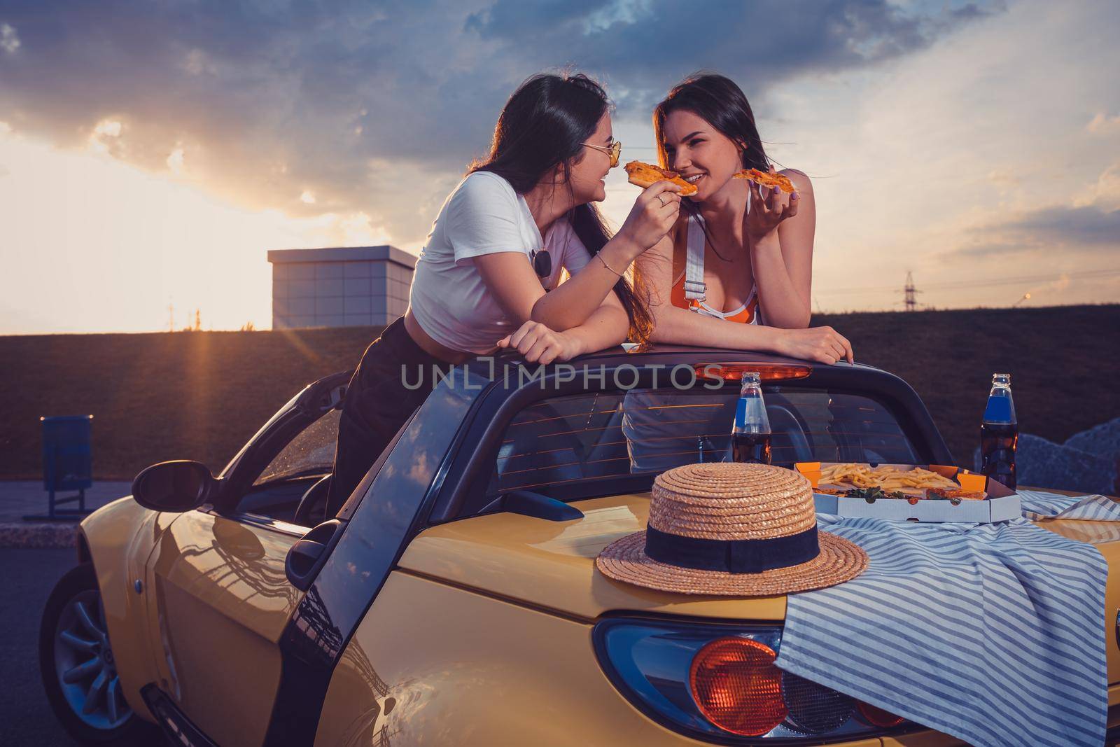 Two young females eating pizza, smiling, posing in yellow car with french fries, hat and soda in glass bottle on its trunk. Fast food. Mock up by nazarovsergey