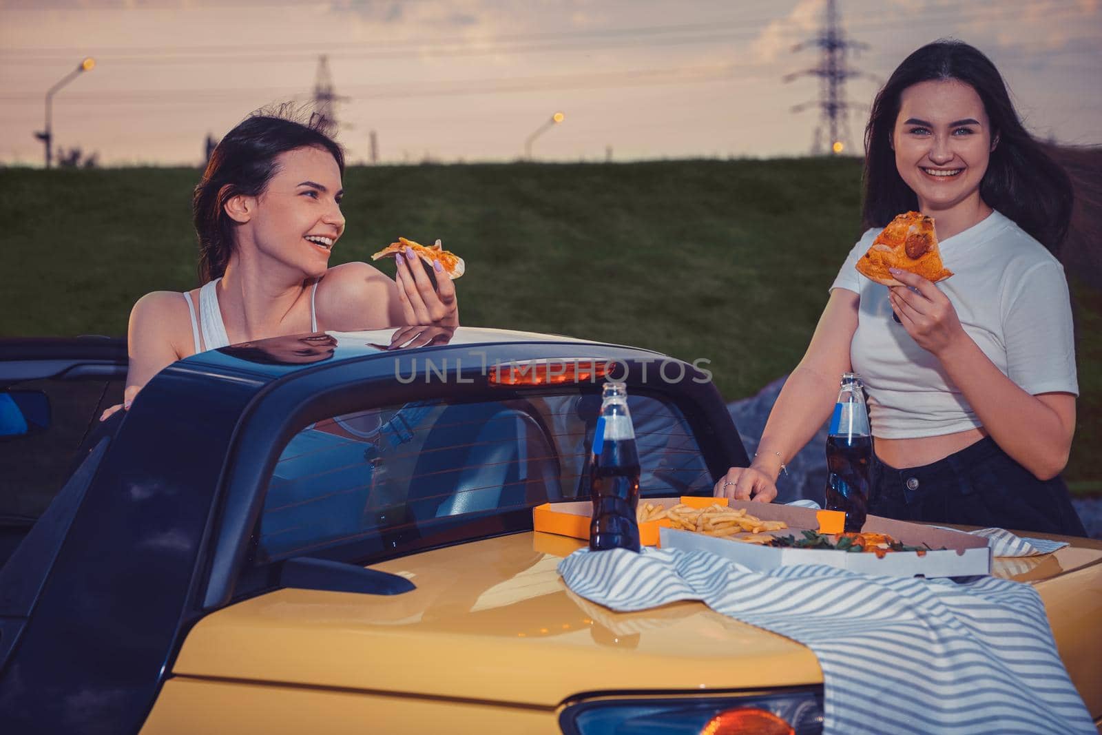Pretty ladies in casual clothes smiling, eating pizza, posing in yellow car cabrio with french fries and soda in glass bottles on its trunk. Fast food. Summer evening. Close up, copy space, mock up