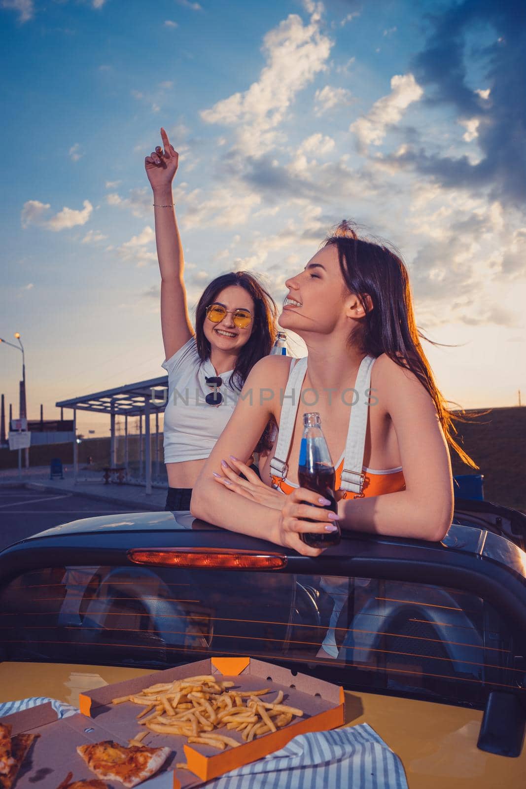 Happy females in casual clothes smiling, having fun and drinking soda in glass bottles, posing in yellow car with french fries and pizza on its trunk. Fast food. Summer evening. Close up, copy space