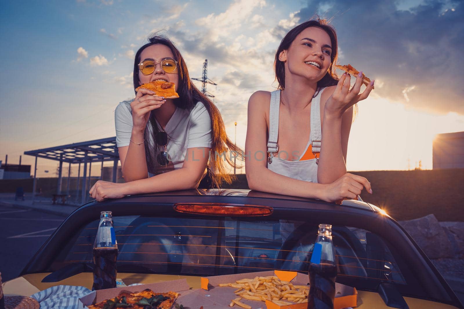Charming women are eating pizza and smiling while posing in yellow car with french fries and soda in glass bottles on trunk. Fast food. Copy space by nazarovsergey