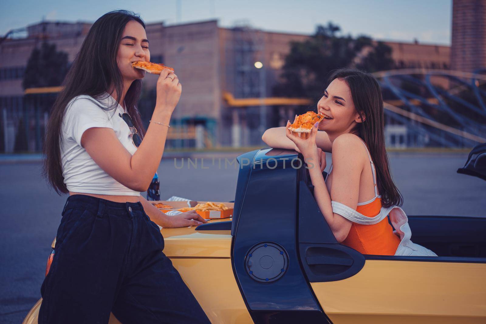 Pretty women in casual outfit are eating pizza, posing in yellow car cabrio with french fries and soda in glass bottle on its trunk. Fast food. Industrial zone. Summer evening. Close up, copy space