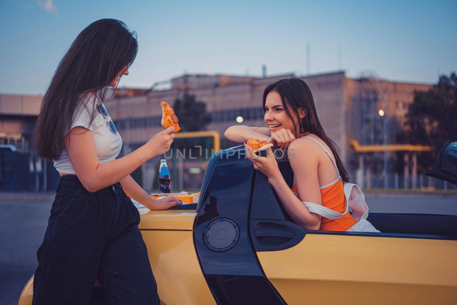 Pretty young girls in casual outfit are smiling, eating pizza, posing in yellow car cabrio with french fries and soda in glass bottle on its trunk. Fast food. Industrial zone. Close up, copy space