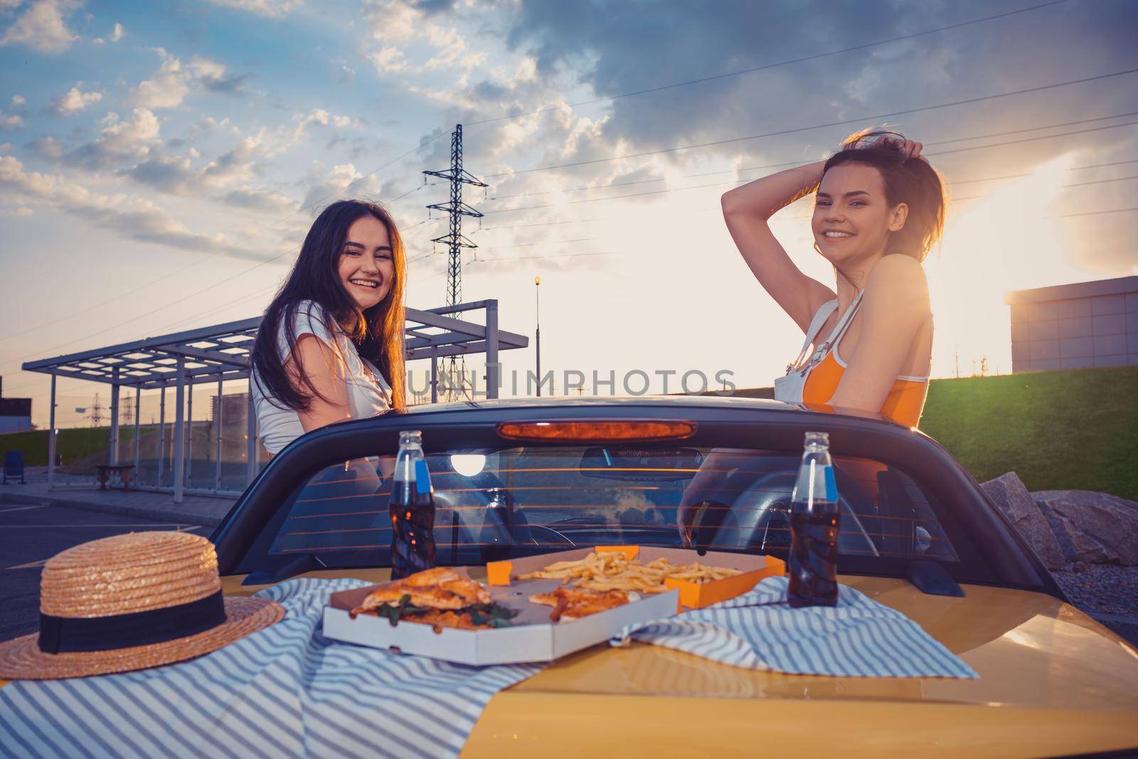 Young friends females in casual clothes are smiling, posing in yellow car cabrio with french fries, pizza and soda in glass bottles on its trunk. Fast food. Summer evening. Copy space, mock up